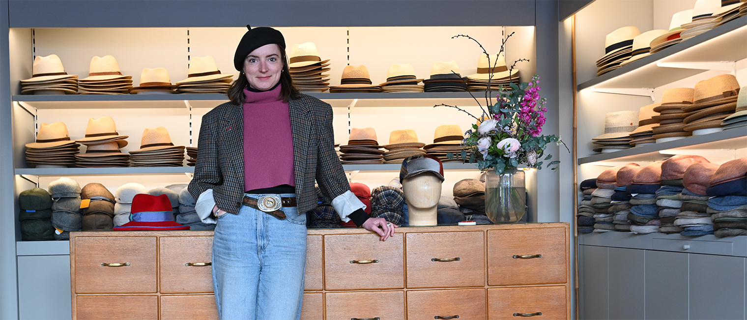 Modistin Laura Zieger in ihrem Geschäft in Würzburg vor einer Kommode, auf der Hüte liegen; im Hintergrund Regale mit Hüten.