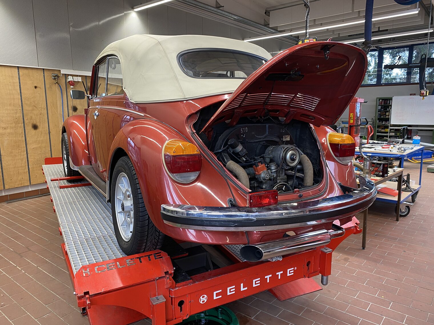 Ein roter VW Käfer mit offenem Motorraum steht in einer Werkstatt auf einer Hebebühne.