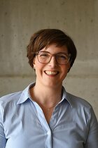 Frau in hellblauer Bluse mit kurzen braunen Haaren lächelt in die Kamera