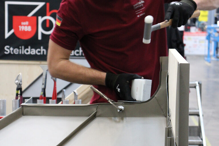 Spengler klopft an Blech mit Hammer an einen Dachmodell